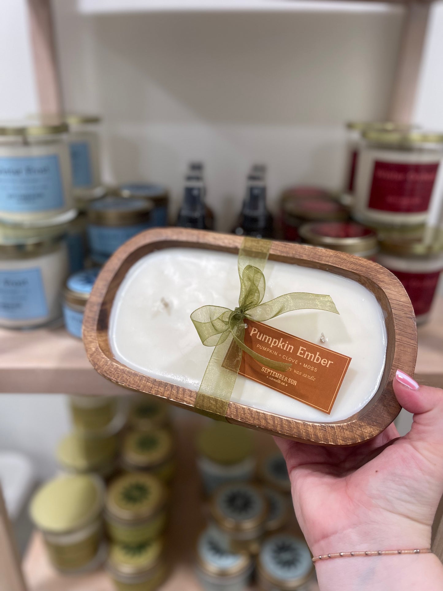 Hand holding a scented candle labeled 'Pumpkin Ember' with a blurred store shelf in the background.